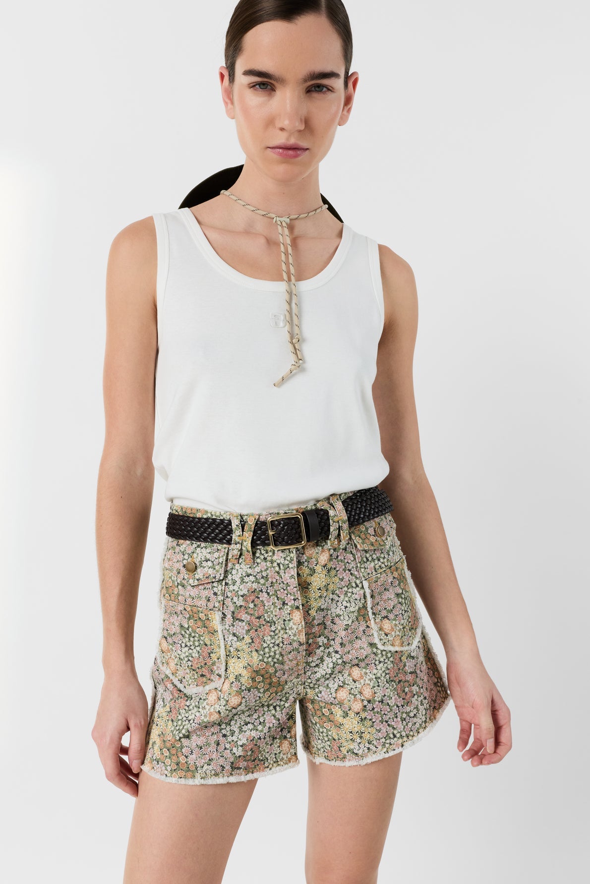 A woman wears a white sleeveless top, motif floral high-waisted shorts with a black belt, and a rope-like necklace. She stands facing forward against a plain white background.