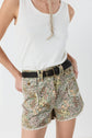 Woman wearing a white sleeveless top and high-waisted floral shorts (motif floral, taille haute) with a black woven belt, standing with one hand on her hip against a plain background.