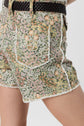 Person wearing high-waisted floral shorts with frayed edges, white trim, and a brown braided belt. These taille haute short en jean feature a large back pocket and a colorful motif floral in shades of green, peach, yellow, and white.