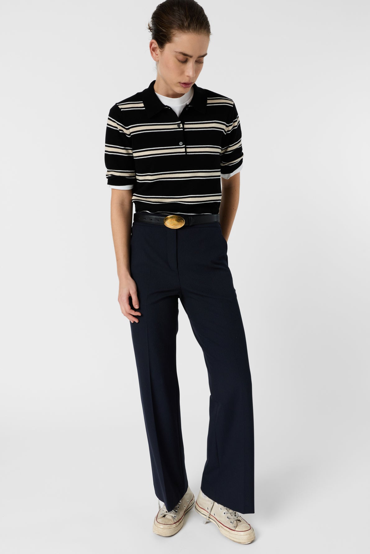 Person wearing a black and white striped polo shirt, dark wide-leg trousers, a wide black belt with a large round buckle, and white sneakers, standing against a plain white background.