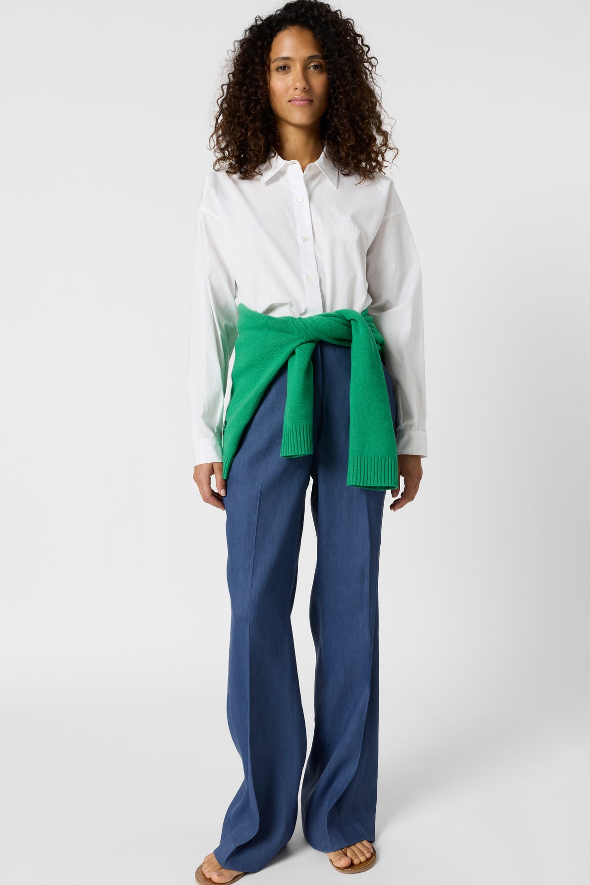 A woman with curly hair wears a white button-up shirt, blue wide-leg pants, sandals, and a green sweater tied around her waist. She stands facing forward against a plain white background.