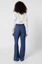 A person with curly hair, wearing a white long-sleeve shirt and blue wide-leg pants, stands facing away from the camera against a plain white background.