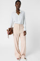 A woman stands wearing a white blouse, light beige high-waisted trousers, and white loafers, holding a brown handbag. She has short hair and layered gold necklaces. The background is plain white.