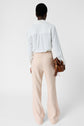 A person stands facing away, wearing a white long-sleeve blouse and loose beige pants, holding a brown handbag with tassel details against a plain white background.