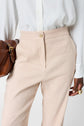 A person wearing a white button-up shirt and beige high-waisted trousers holds a brown leather bag and wears gold rings on their fingers.