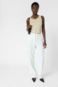A person stands against a white background, wearing a beige sleeveless top, elegant pantalon microfibre blanc tailored for comfort and style, pointed black shoes, and gold jewelry, holding a small black handbag.