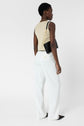 A person stands against a white background, wearing a beige sleeveless top, a white pantalon femme wide-leg style made from soft microfibre, and black pointed shoes, holding a black shoulder bag with a gold chain strap.