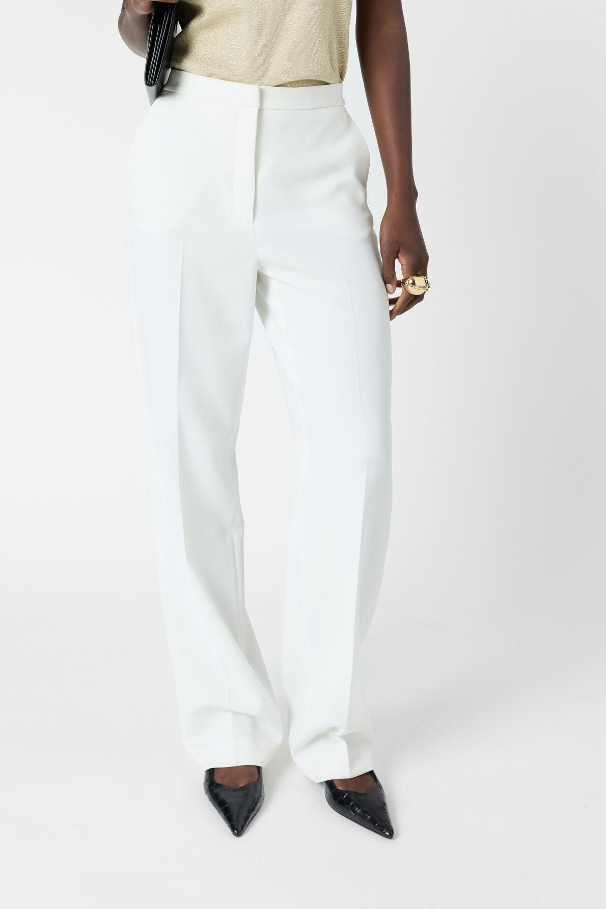 A person wearing high-waisted white pantalon femme in microfibre, paired with a light-colored top and black pointed-toe heels, stands against a white background holding a black clutch. Only the torso and legs are visible.