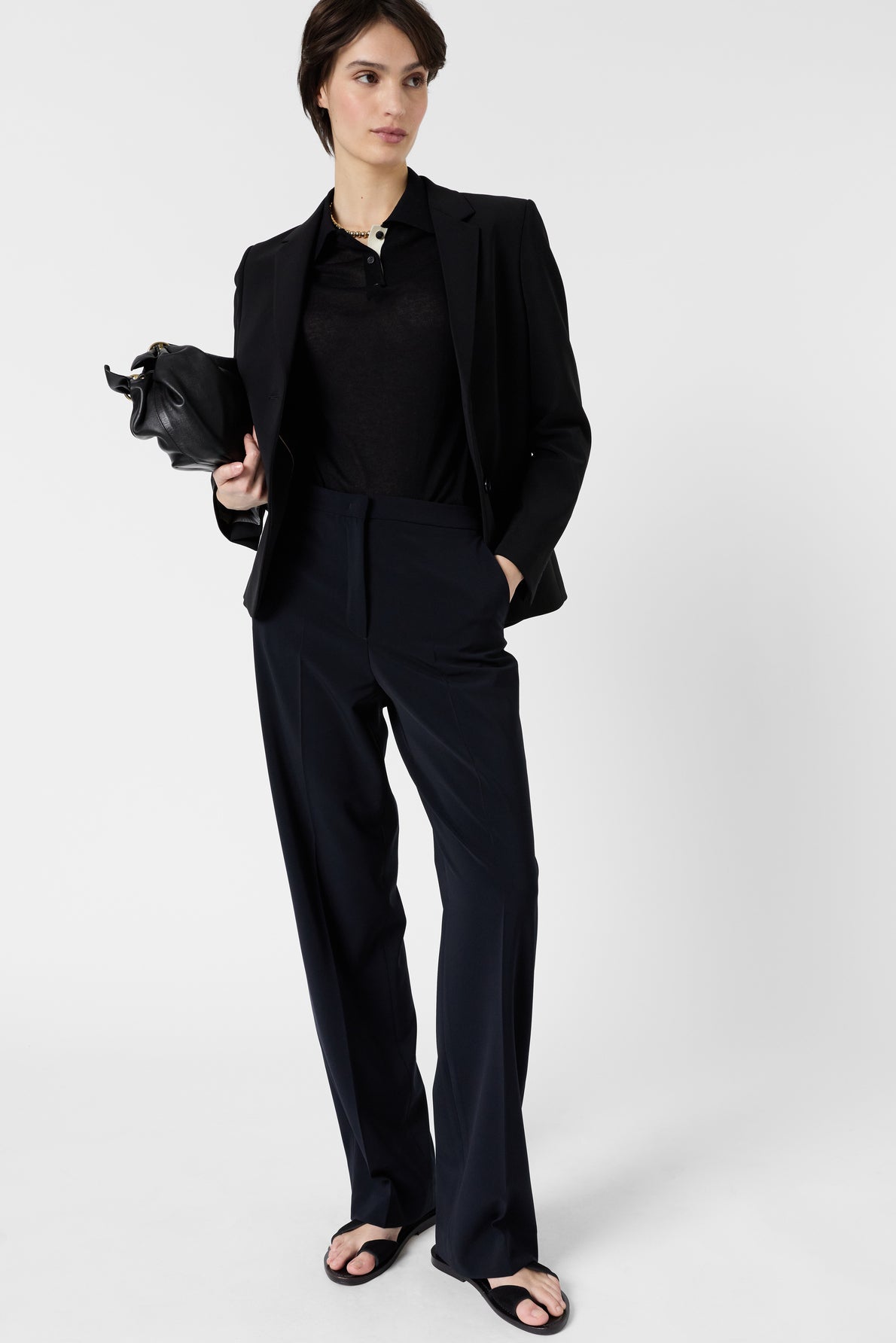 A person wearing a black blazer, black top, pantalon droit noir, and black sandals stands holding a small black bag with one hand, with the other hand in their pocket, against a plain white background.