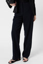 Person wearing black pantalon droit, black blazer, and black top, holding a black bag, with black sandals on their feet. The background is plain white. The person’s face is not visible.