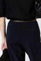A person wearing a pantalon bleu marine styled as a pantalon droit and a black top is seen from behind, holding a black item in their left hand. The waistband of the trousers has a subtle metallic sheen.
