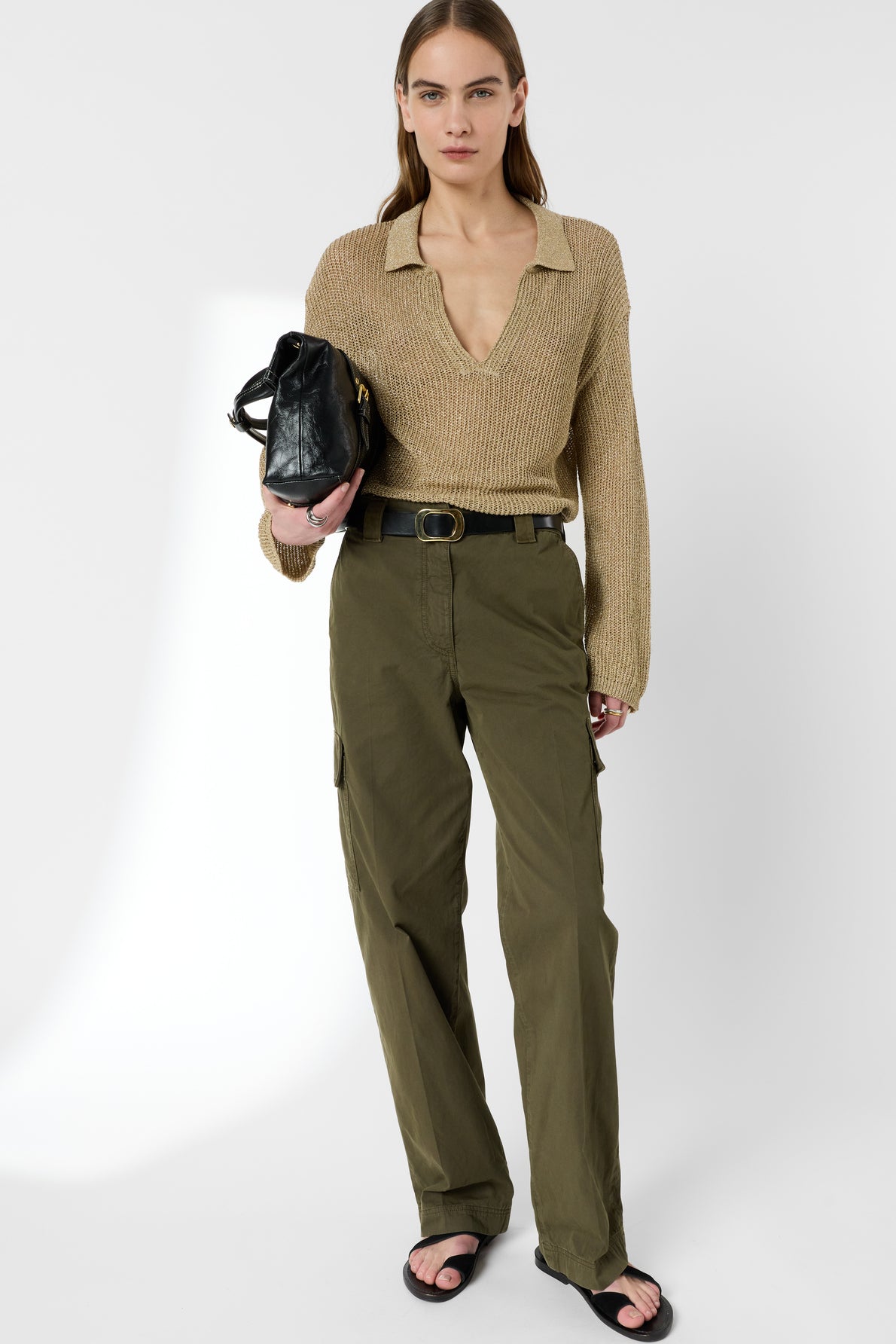 A woman stands against a white background, wearing an open-collar beige sweater and a pantalon militaire femme in olive green, featuring grandes poches and a stylish belt. She pairs the look with black sandals, holds a black handbag under her arm, and has straight hair.