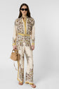 Woman wearing sunglasses, a cream-colored matching set with gold and floral patterns, holding a tan woven handbag, and wearing flat sandals, stands against a plain light background.