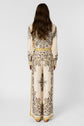 A person with long wavy brown hair stands facing away, wearing a cream-colored outfit with intricate floral patterns and yellow accents, against a plain light background.
