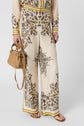 A person wearing cream-colored, wide-leg pants with an intricate floral pattern, a matching long-sleeve top, tan sandals, and holding a woven tan handbag.