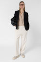 A woman wearing glasses, a black blazer, a patterned blouse, loose white coton pants, and white sneakers holds a black handbag with a subtle logo GD and stands against a plain white background.