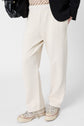 Person wearing cream-colored coton pantalon de jogging with a subtle logo GD, beige graphic shirt, black blazer, and white sneakers, holding a black bag. Only the lower half of the outfit is visible against a plain background.