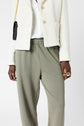 A person wearing a white textured jacket, white shirt, and an olive green pantalon de jogging 100% coton stands against a plain background. They hold a black purse with a gold chain and wear gold rings, showcasing un look kaki tendance.