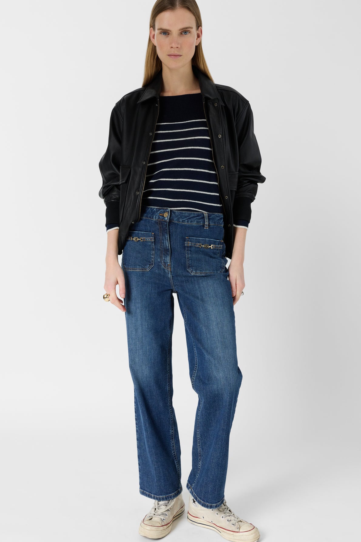A woman stands wearing a black jacket, navy and white striped sweater, blue jeans coupe droite with front poches monogrammes dorés, and white sneakers against a plain white background.