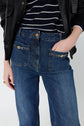 Person wearing a jean brut à coupe droite with poches monogrammes dorés on the pockets, a black and white striped sweater, and a black leather jacket. One hand is in a front pocket. The image is cropped at the midsection.