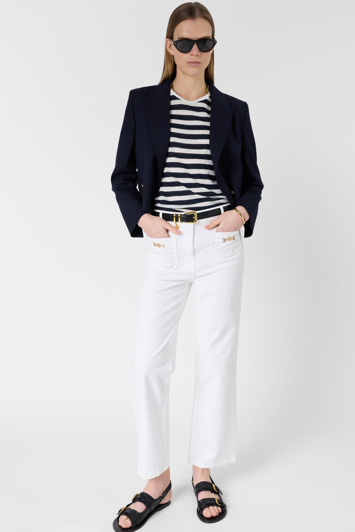 Woman wearing black sunglasses, navy blazer, black and white striped shirt, and a coupe droite jean blanc with poche monogramme stands against a plain white background with hands in her pockets. She completes the look with a black belt featuring a gold buckle and stylish black sandals.