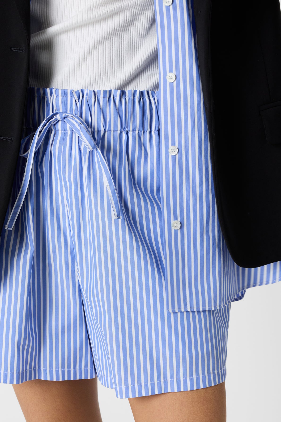 A person wearing a white ribbed top, blue and white vertical striped shorts with a drawstring, and a dark jacket.