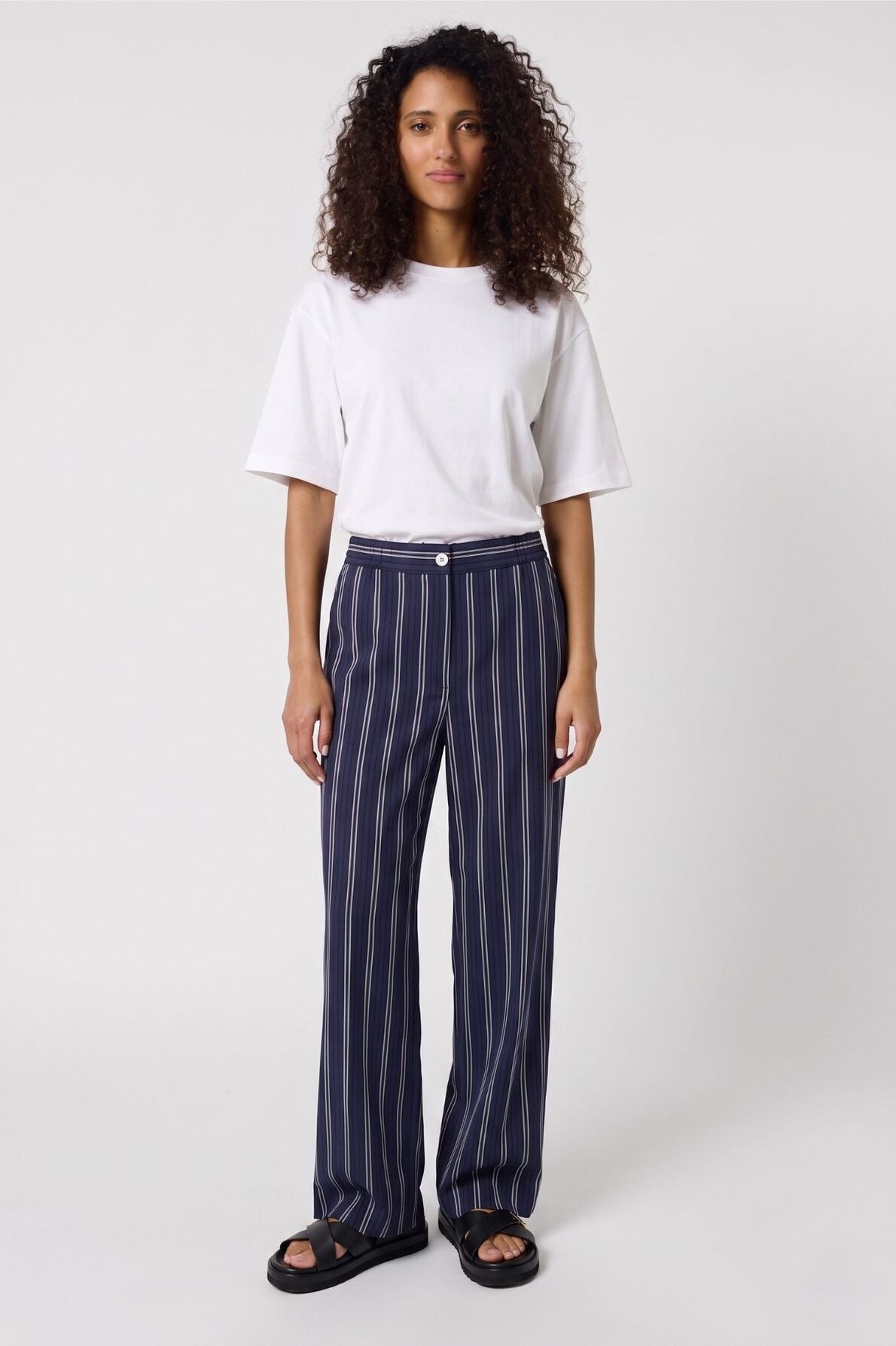 Woman with curly hair wearing a white t-shirt, navy blue pants with white vertical stripes, and black sandals, standing against a plain light background.