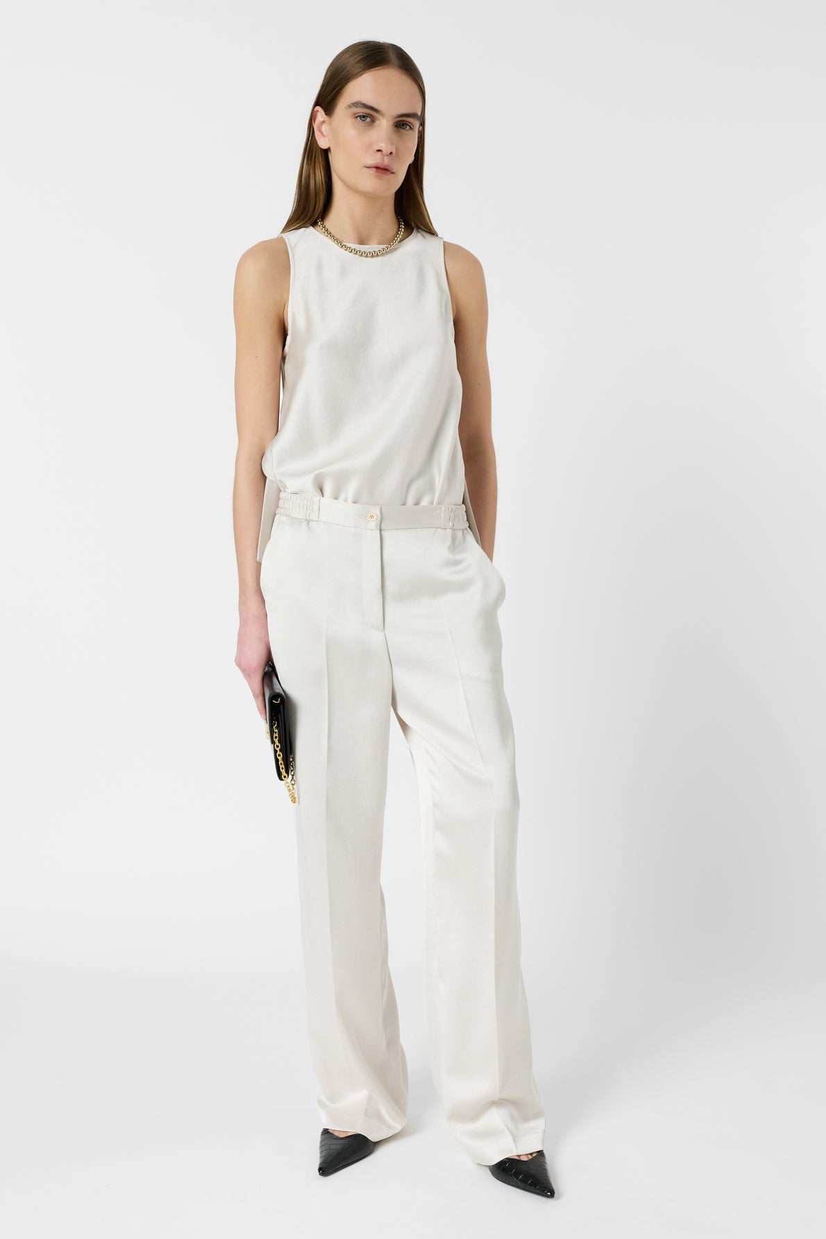 A woman stands against a white background wearing a sleeveless white top, white tailored pants, black pointed shoes, and holding a black clutch.