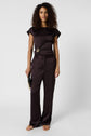 A woman with curly hair wears a dark brown satin top with a gold buckle and matching pantalon fluide marron, holding a black clutch, standing against a plain white background.
