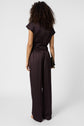 Person with curly hair wearing a loose-fitting, short-sleeve, dark brown satin jumpsuit featuring a pantalon fluide silhouette stands facing away, holding a black clutch against a plain white background.