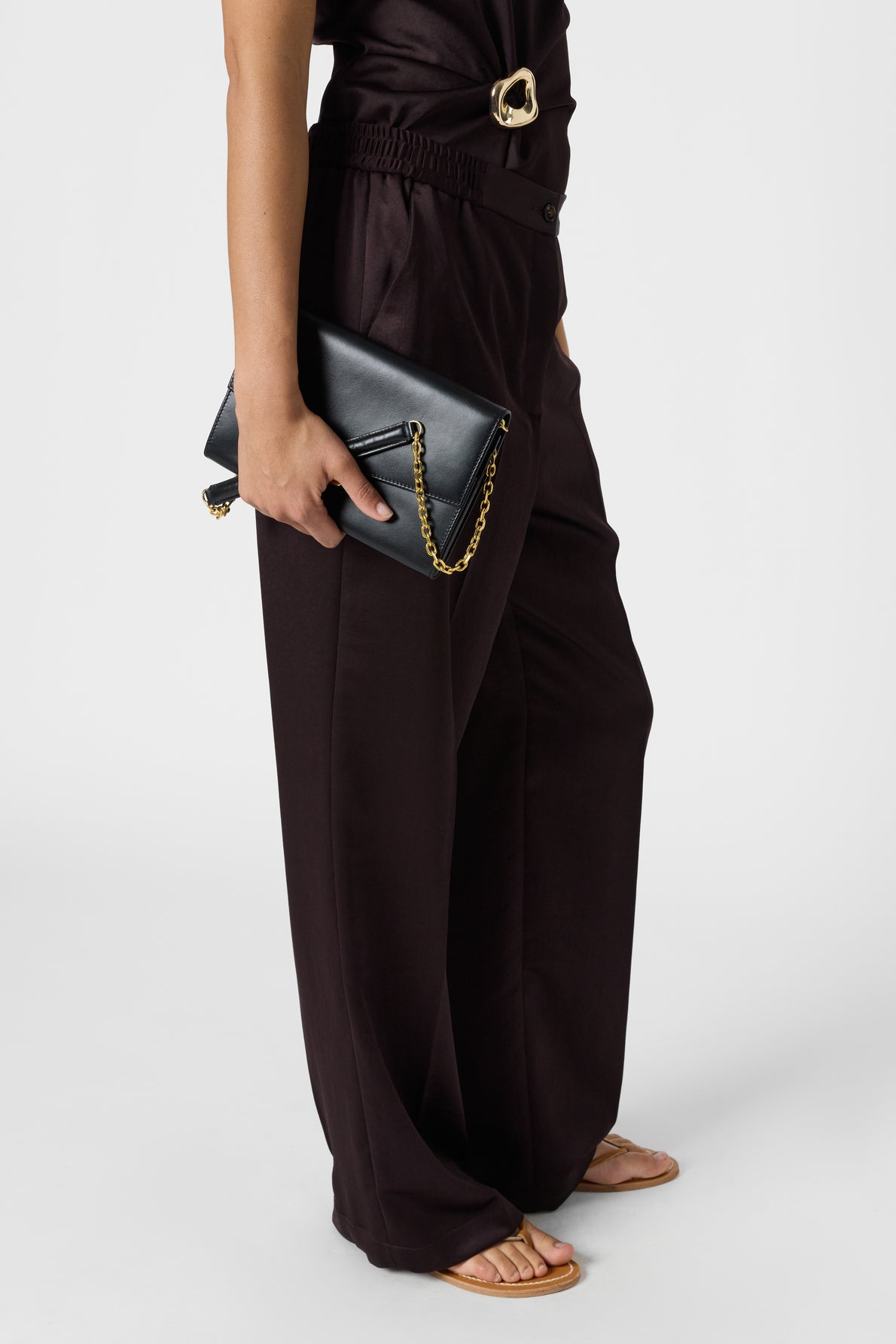Person standing in a black pantalon fluide, holding a black clutch with a gold chain. They are wearing sandals and a black top with a gold buckle detail. Background is plain white.