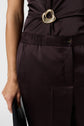 A person wearing a pantalon marron and a matching top with a gold decorative buckle, holding a black object in their left hand. Only the lower torso and part of the left arm are visible.