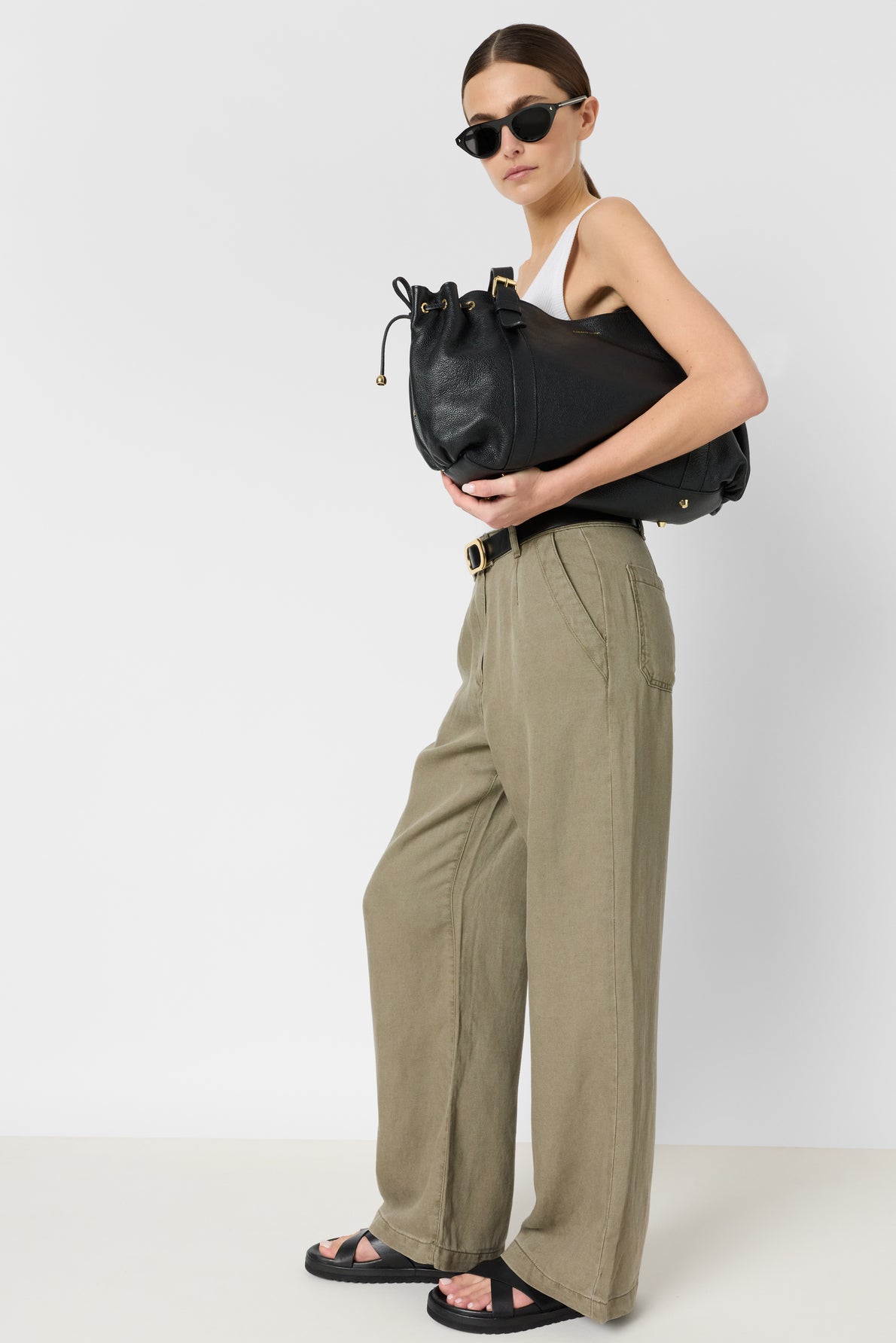 Woman wearing sunglasses, a white tank top, and a chic pantalon coupe droite in olive green lyocell-linen blend, paired with black sandals. She holds a large black handbag under her arm, standing confidently against a plain white background—an effortlessly stylish look with the perfect pantalon kaki femme.