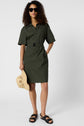 A woman wearing sunglasses and a dark green belted dress stands with one hand in her pocket, holding a woven hat in the other. She wears black sandals and stands against a plain background.