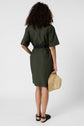 A person with curly hair, wearing a green dress and black sandals, stands facing away, holding a straw hat in their right hand against a plain white background.
