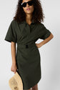 Woman in dark green belted shirt dress wearing black sunglasses, holding a straw hat, standing against a plain light background.