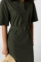 A person wearing a belted, short-sleeved, dark green dress holds a woven straw bag. Their face is not visible; the focus is on the dress and accessories.