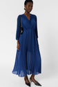 A woman wearing a long-sleeved, pleated, deep blue midi dress, black pointed flats, gold hoop earrings, and a black shoulder bag stands against a plain white background.