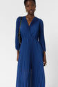 Woman wearing a long-sleeved, deep blue, pleated dress with a V-neckline. She carries a black shoulder bag with a gold chain and wears gold hoop earrings. Background is plain white.