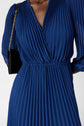 A person wearing a blue, pleated, V-neck dress with long sleeves and holding a black handbag with a gold chain strap. The image is cropped at the shoulders and mid-thigh.