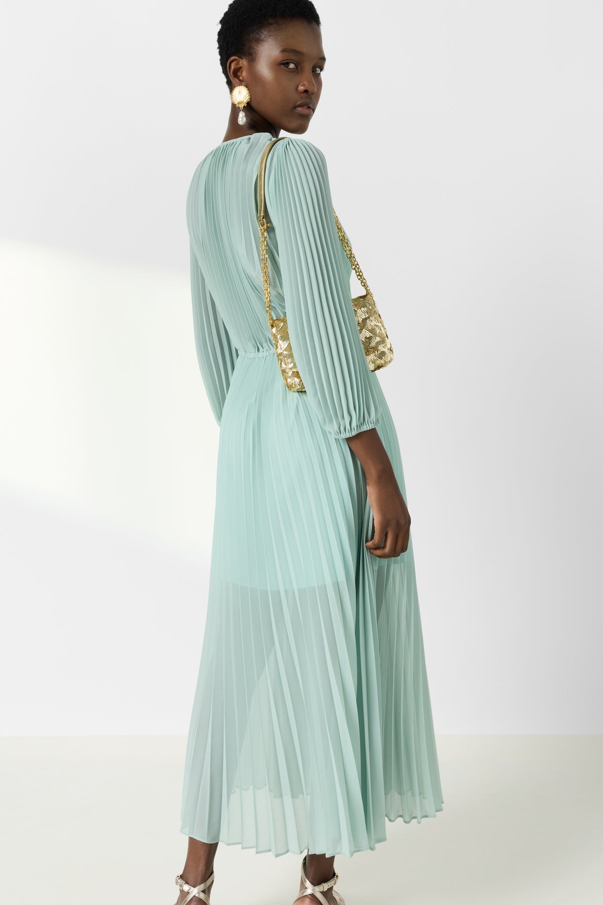 Woman in a light blue pleated long-sleeve dress stands with her back turned, looking over her shoulder. She carries a gold sequin bag and wears dangling earrings and heeled sandals.