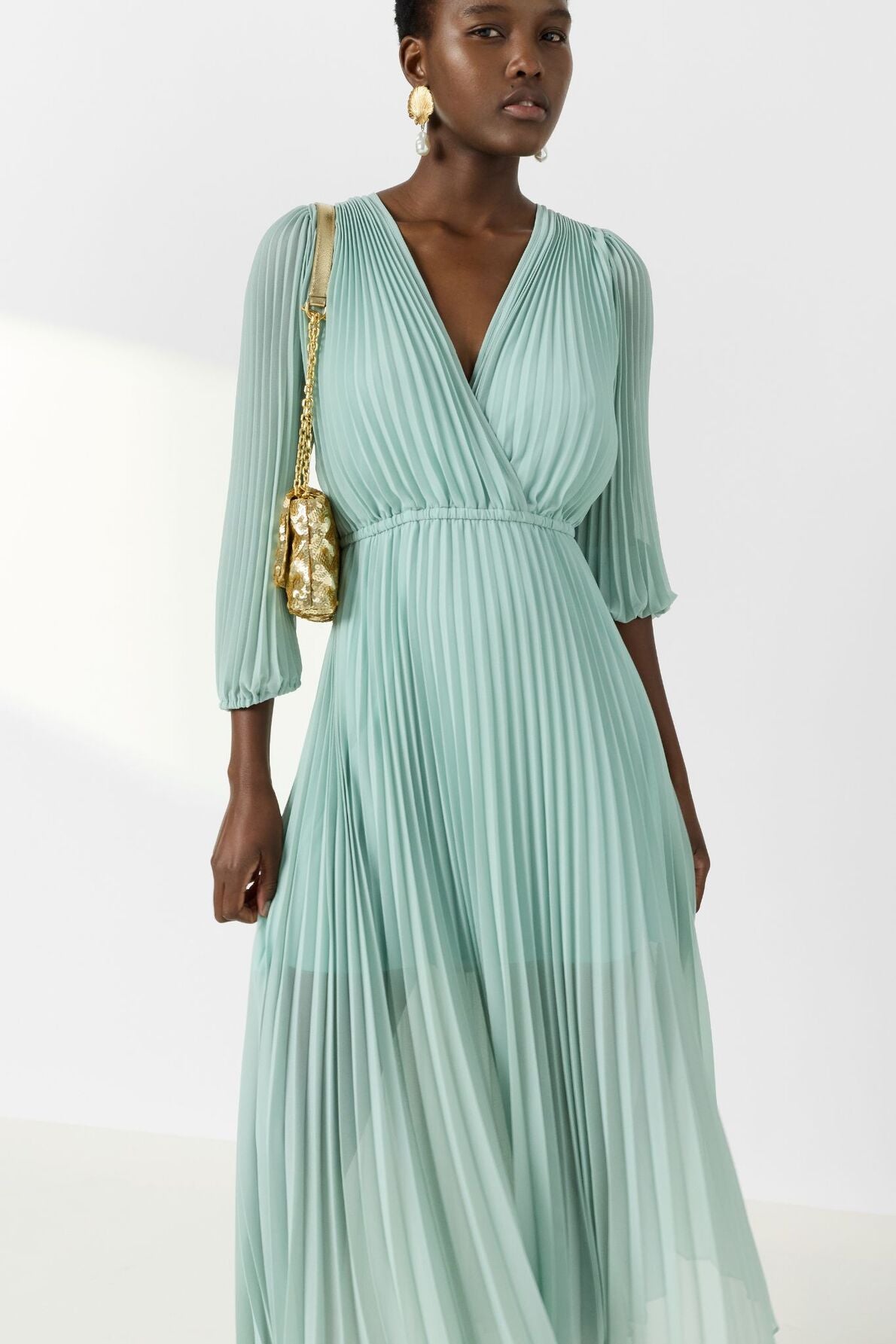 A woman wearing a light green, pleated, long-sleeved, V-neck dress and gold earrings carries a small gold shoulder bag. She stands against a plain white background.