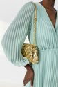 A person in a light green, pleated dress holds a small gold, quilted handbag with a chain strap over their shoulder. The image is cropped to show only the torso and arm.