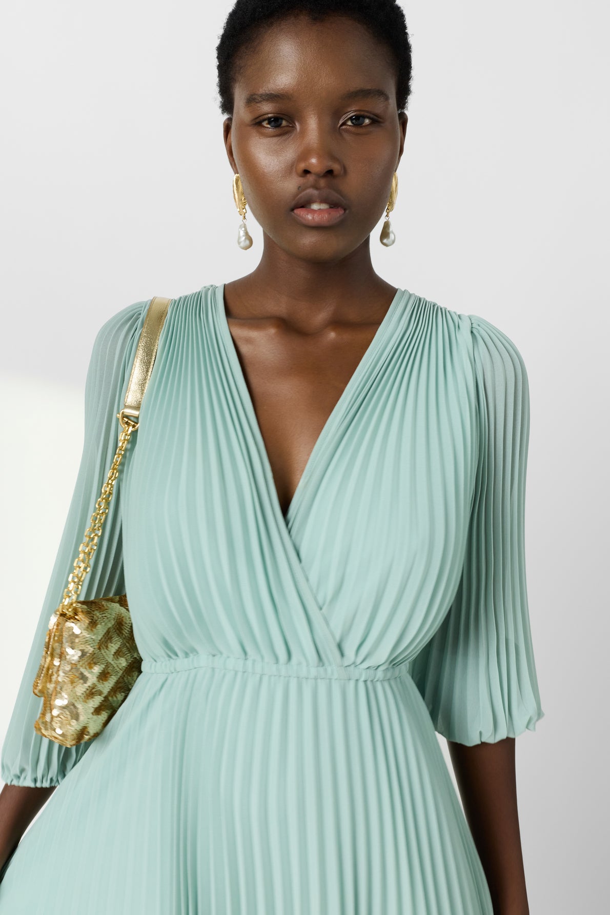 A woman wearing a pleated, light green V-neck dress, gold earrings, and carrying a small gold shoulder bag stands against a plain light background.