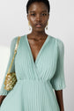A woman wearing a pleated, light green V-neck dress, gold earrings, and carrying a small gold shoulder bag stands against a plain light background.