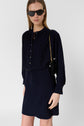Woman wearing a microfibre bleu marine dress with col rond and long sleeves, gold detail buttons, a black handbag with a gold chain, and black sunglasses, standing against a plain light background.