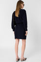 A woman stands facing away, wearing a robe courte navy long-sleeve dress with a col rond, black heeled sandals, sunglasses, and a black shoulder bag with a gold chain. Her straight, light brown hair falls smoothly down her back. The background is plain white.