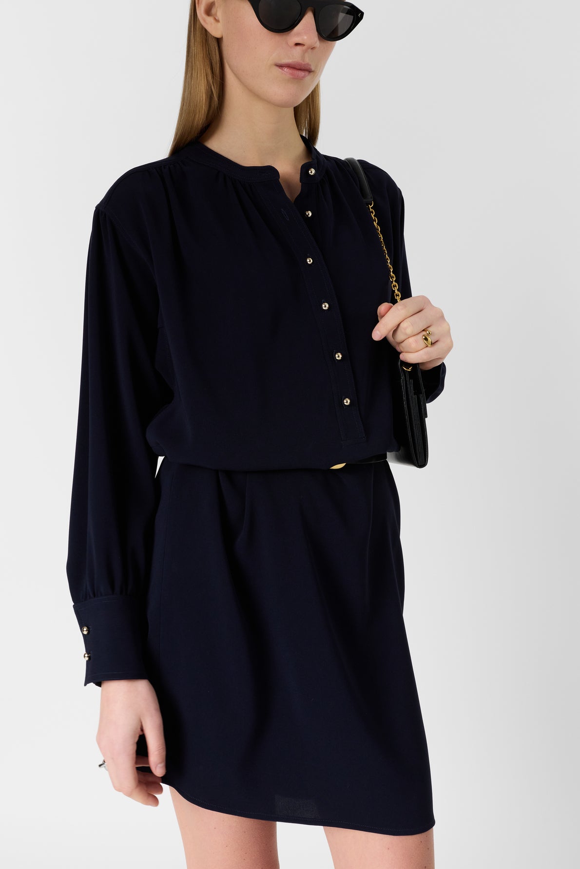 A woman wearing a long-sleeve navy microfibre bleu marine dress with buttons and a col rond, black sunglasses, and a black purse with a gold chain strap stands against a plain white background.