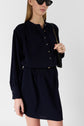 A woman wearing a long-sleeve navy microfibre bleu marine dress with buttons and a col rond, black sunglasses, and a black purse with a gold chain strap stands against a plain white background.