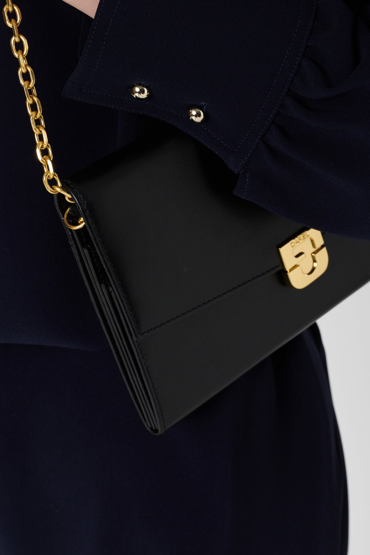 A close-up of a person holding a black leather clutch with a gold chain strap and gold clasp, wearing a microfibre bleu marine long-sleeve garment with silver button details.